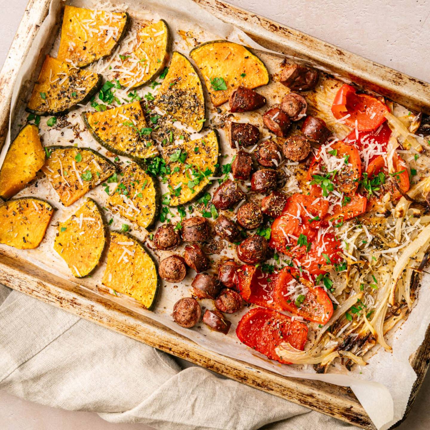 Roasted vegetables on a baking tray with savory sausage. Includes sliced pumpkin, tomatoes, and onions, garnished with herbs and grated cheese. Warm, rustic feel.