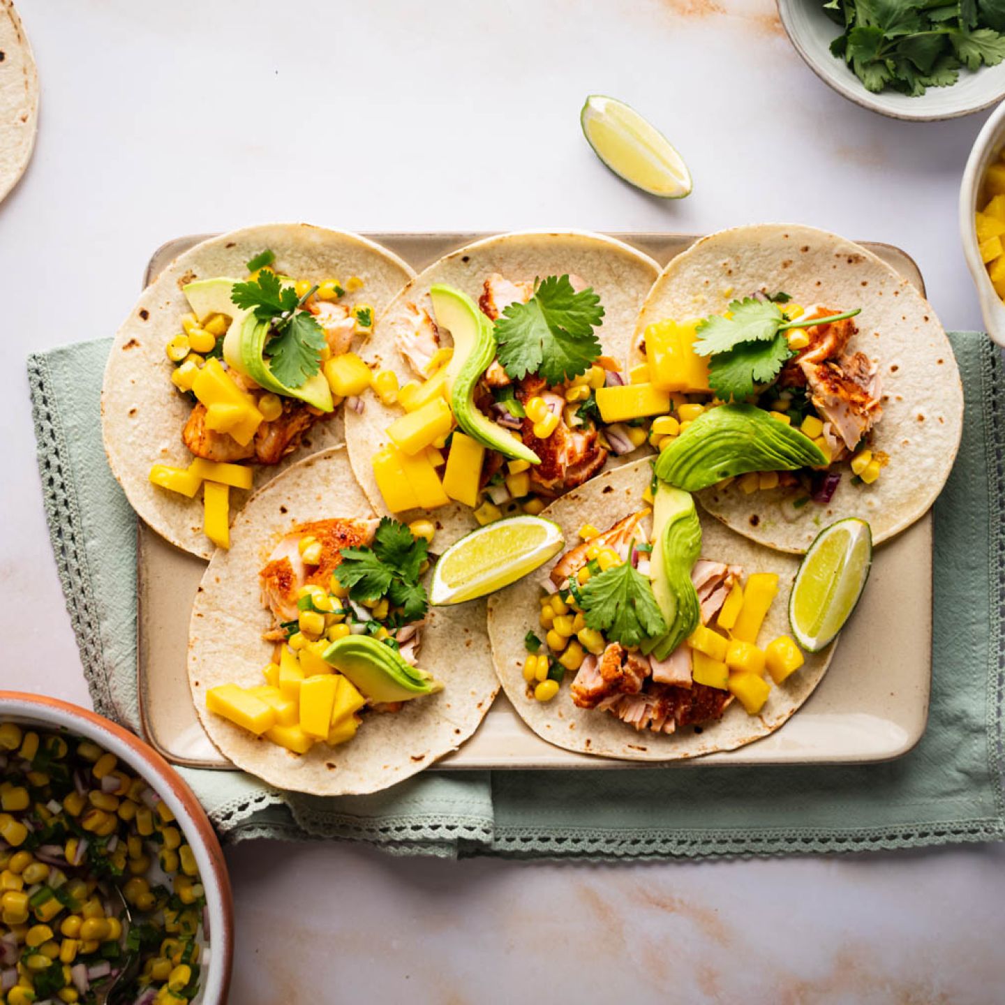 Salmon tacos with corn salsa, mango, fresh avocado slices, and lime wedges on a tray. 