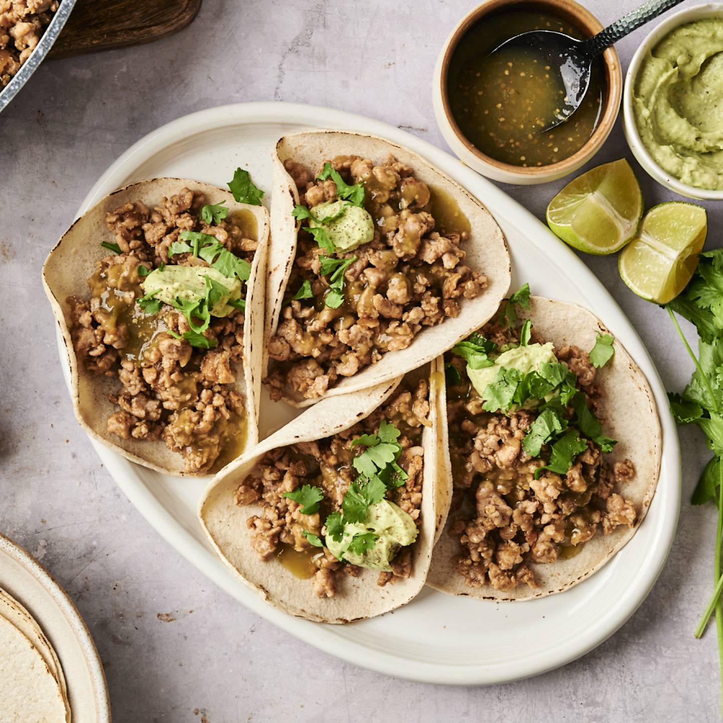 Plate of salsa verde turkey tacos with ground turkey filling, cilantro, and avocado crema, surrounded by lime wedges, salsa verde, and tortillas.