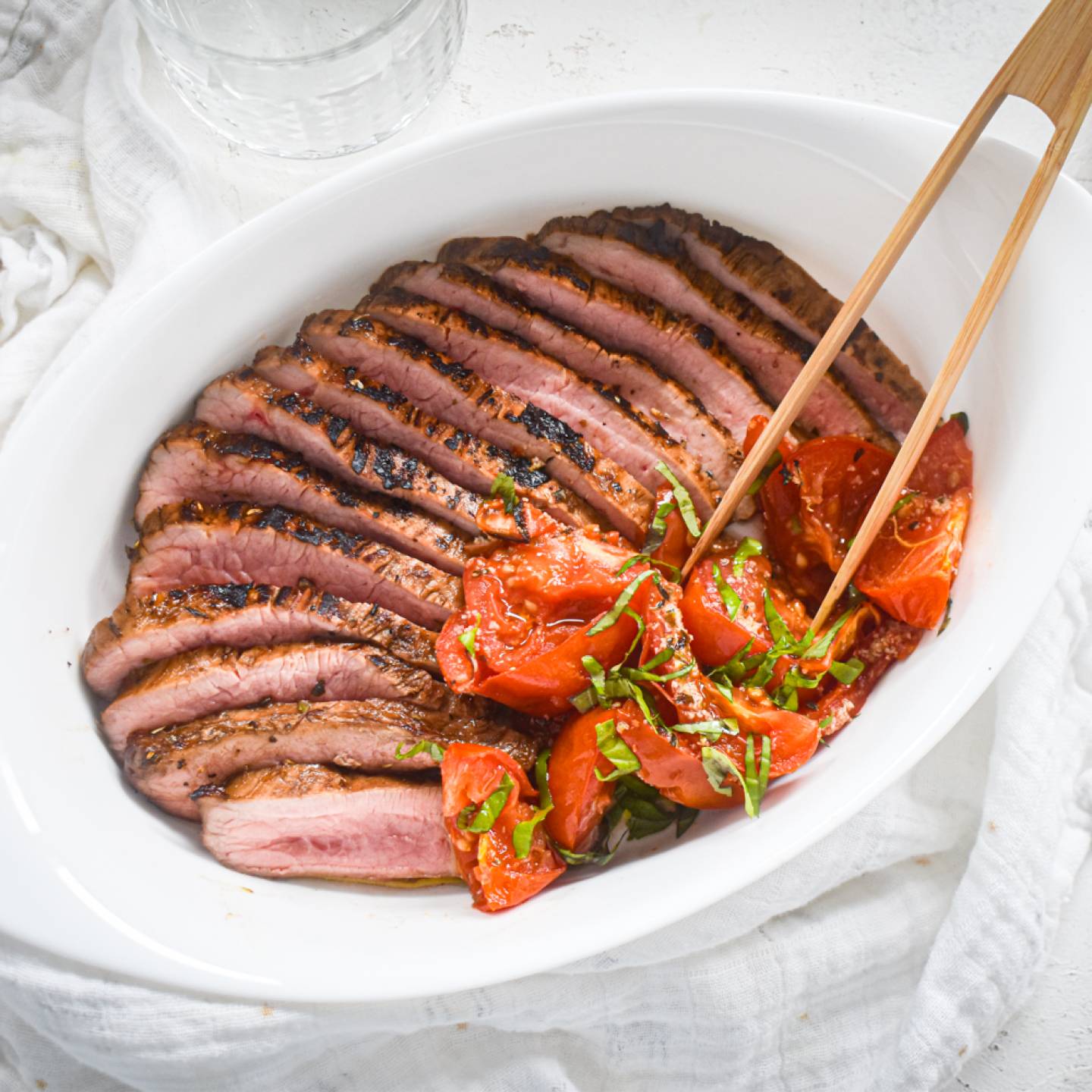 Balsamic flank steak sliced and served with roasted tomatoes in a white plate with wooden tongs.