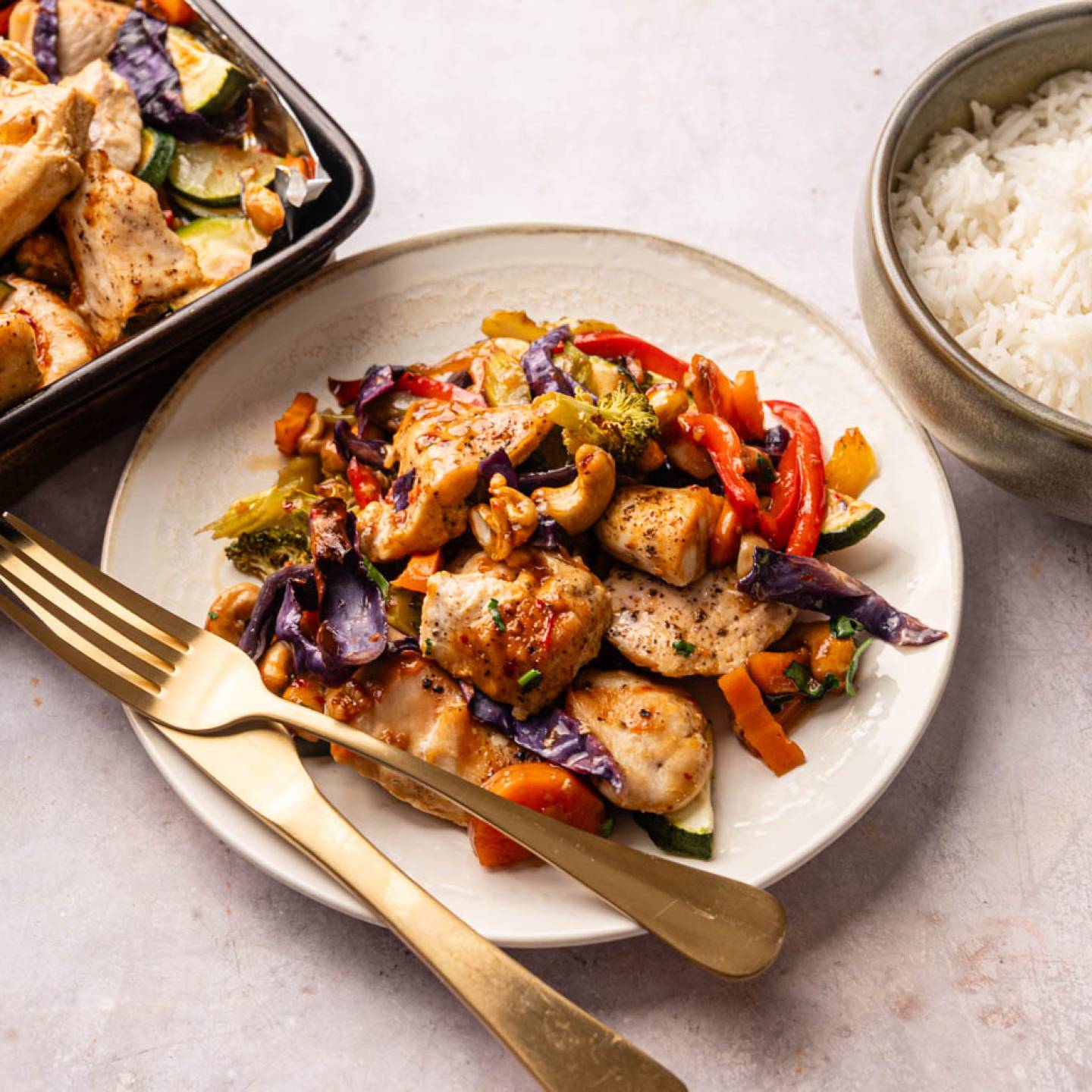 Stir-fried chicken with colorful vegetables, including red peppers and purple cabbage, on a white plate next to a bowl of white rice.
