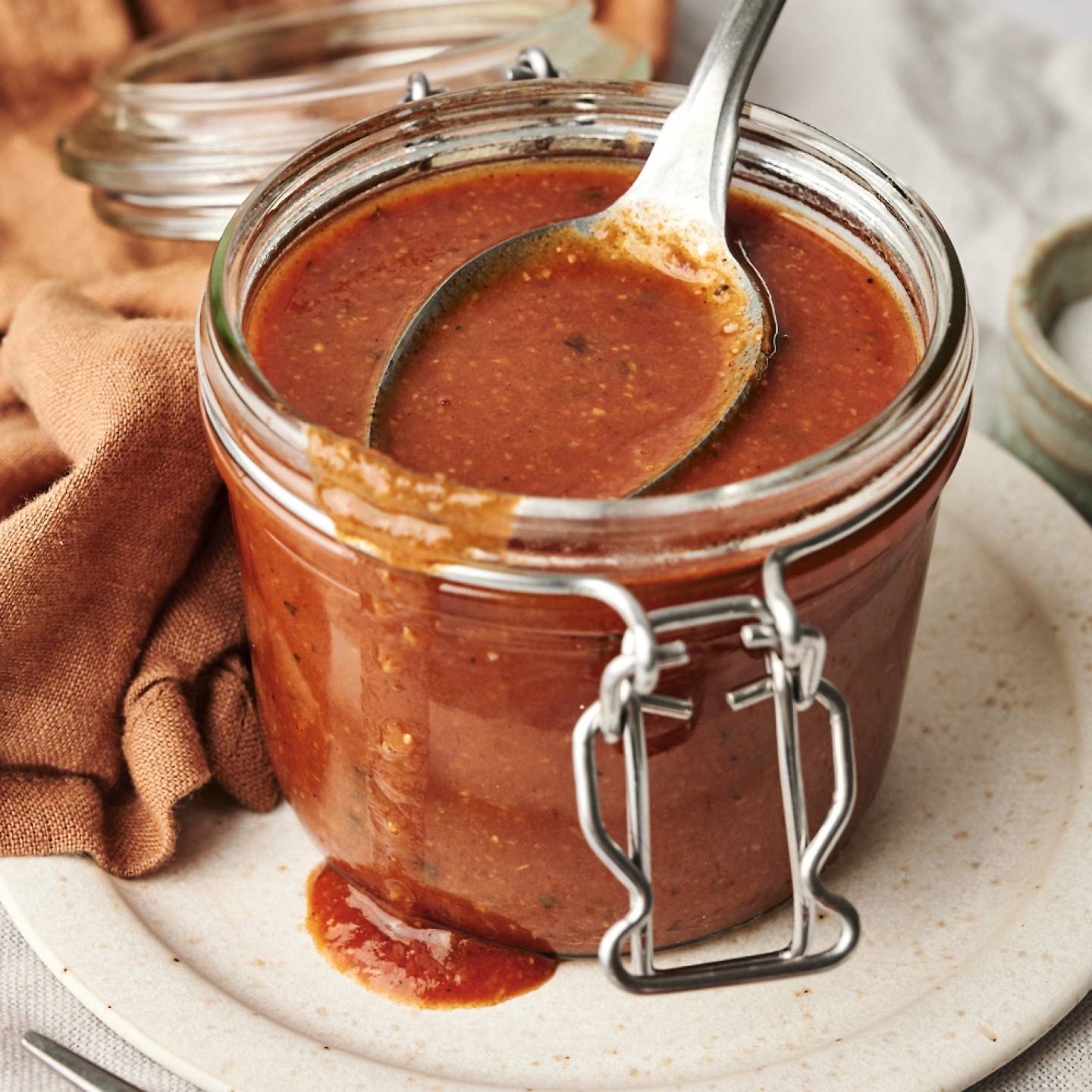 Shortcut Enchilada Sauce A glass jar filled with homemade enchilada sauce, with a spoon resting inside, placed on a ceramic plate with a rustic cloth and a small bowl of salt in the background.
