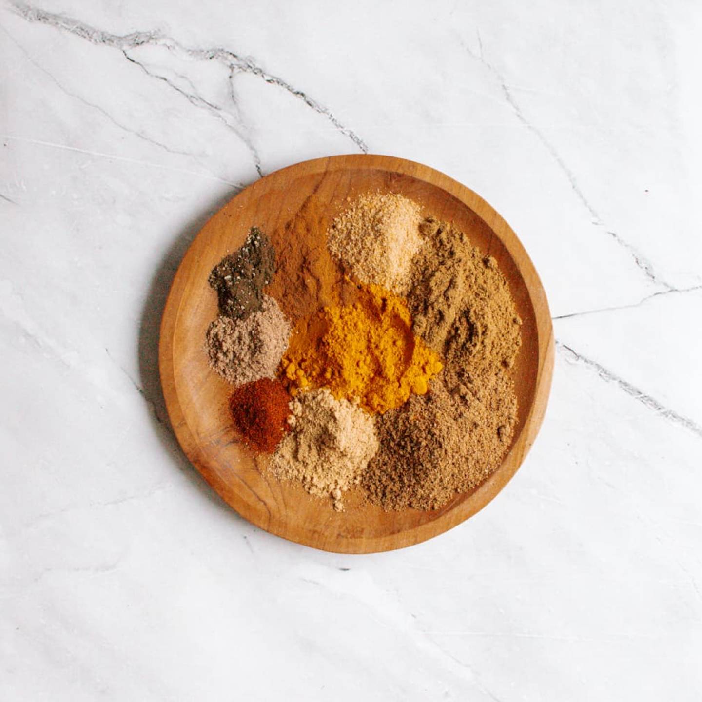 Homemade curry powder on a wooden plate with a variety of ground spices including cumin, coriander, cardamom, ginger, cinnamon, and cayenne.