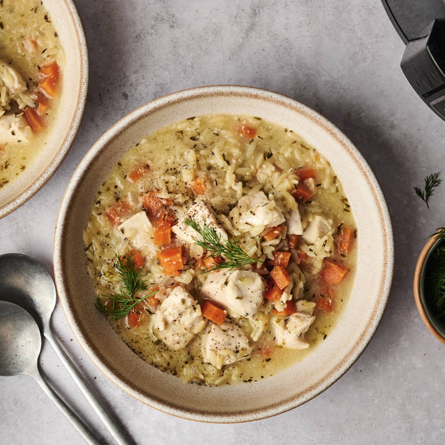 Two bowls of creamy chicken and rice soup garnished with fresh dill and diced tomatoes, accompanied by two spoons and a bowl of herbs.