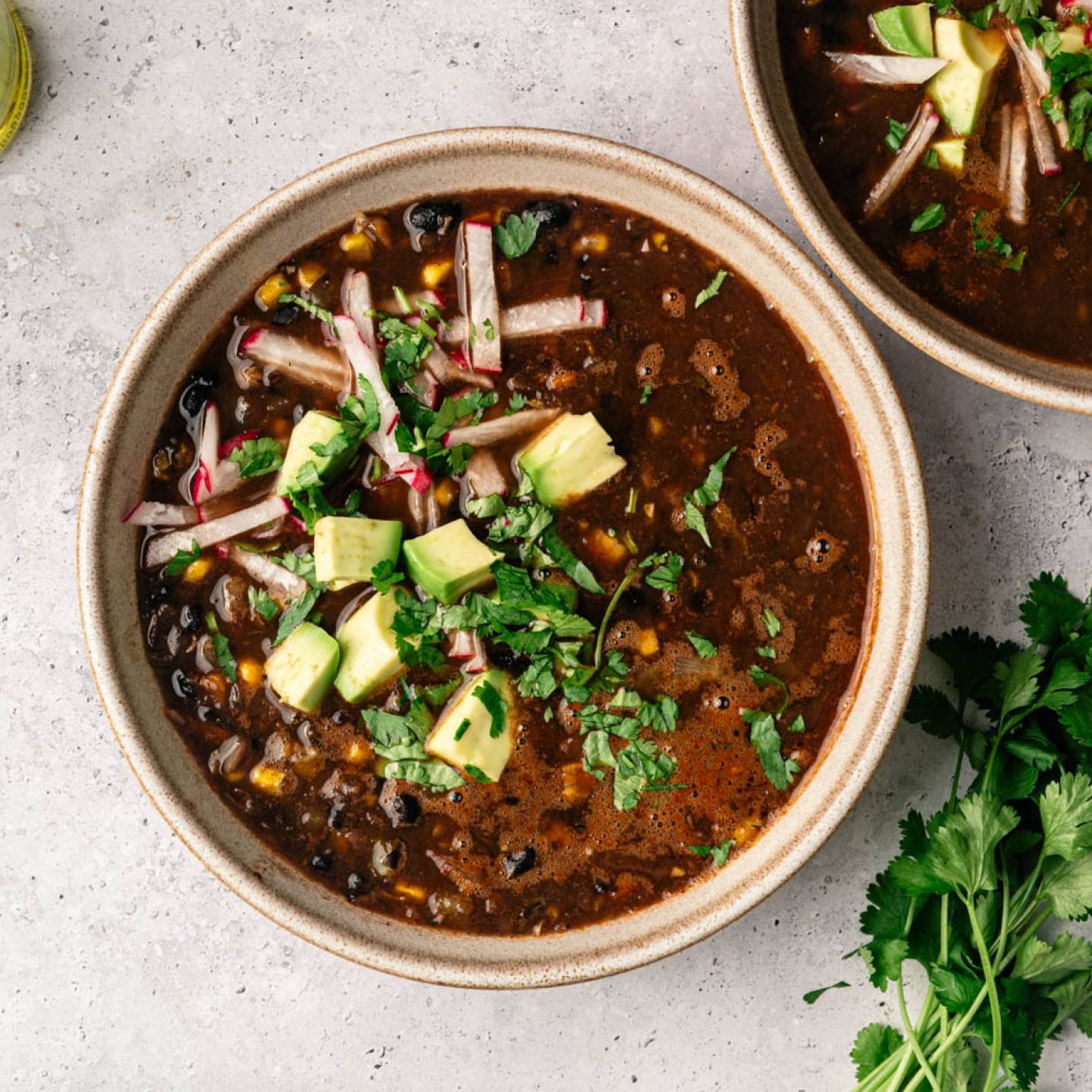 A bowl of black bean soup topped with avocado, cilantro, and radish strips on a textured gray surface. A jar of oil and fresh lime and cilantro are nearby.