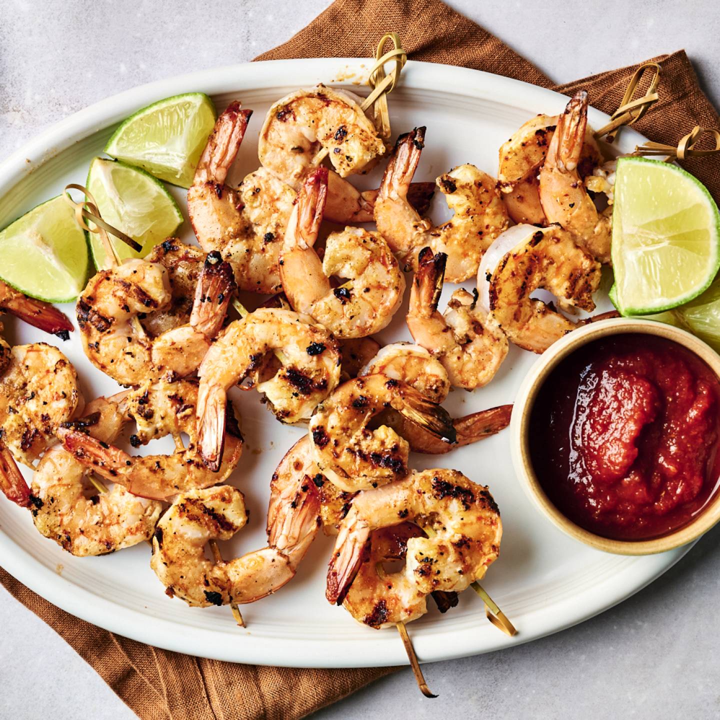 Grilled shrimp skewers served with lime wedges and a bowl of dipping sauce on a white platter over a brown cloth.