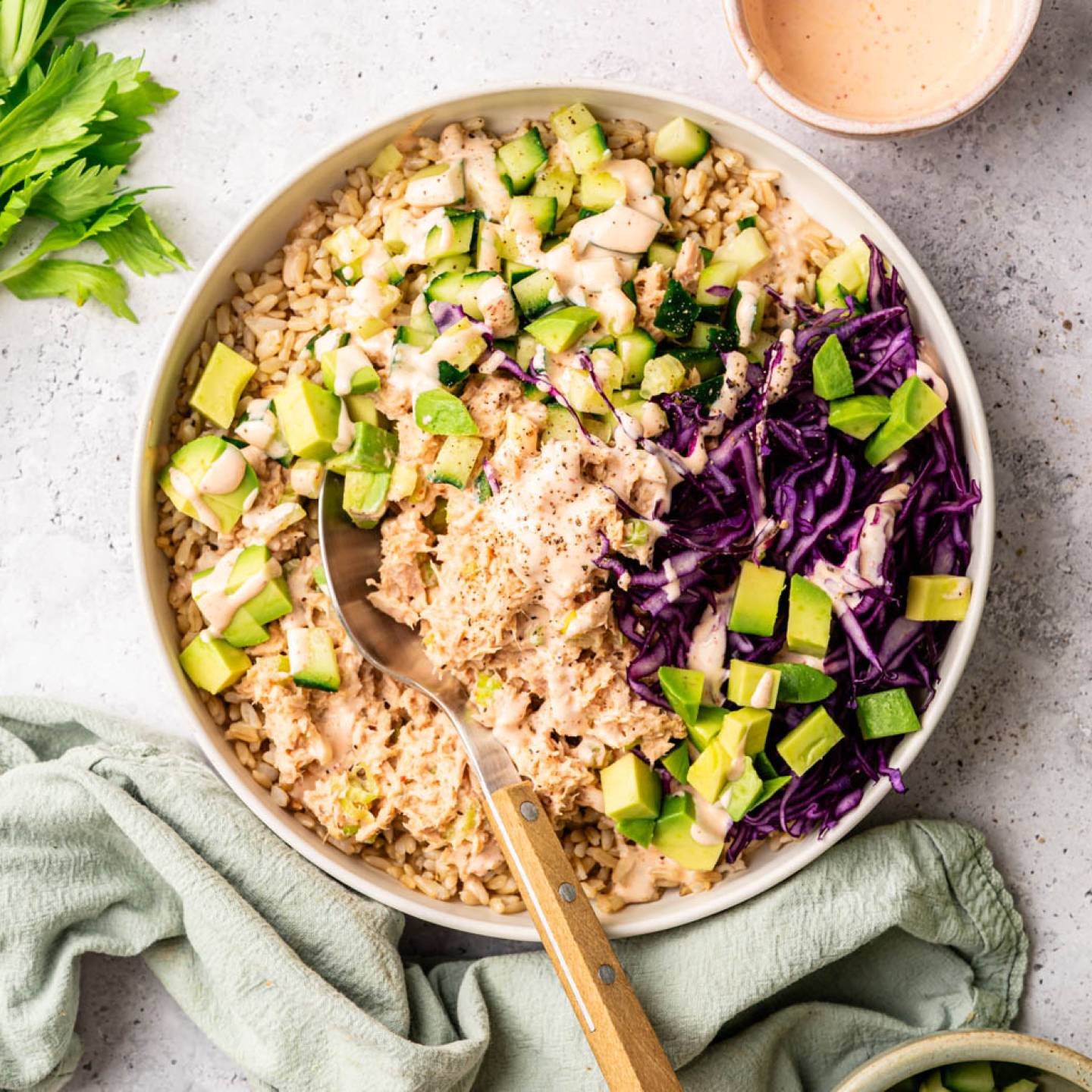 A colorful bowl of brown rice topped with cubed avocado, diced cucumber, purple cabbage, and dressing. Celery and a linen cloth are nearby, creating a fresh and vibrant look.