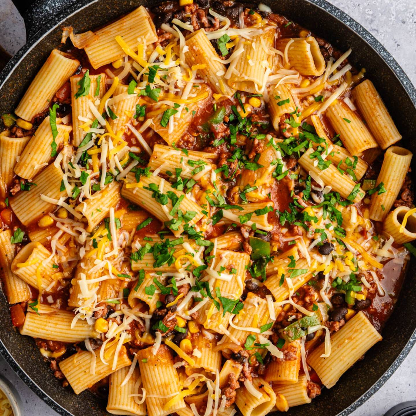 A pan of rigatoni pasta with tomato sauce, ground meat, corn, and black beans, topped with melted cheese and parsley, on a gray surface.