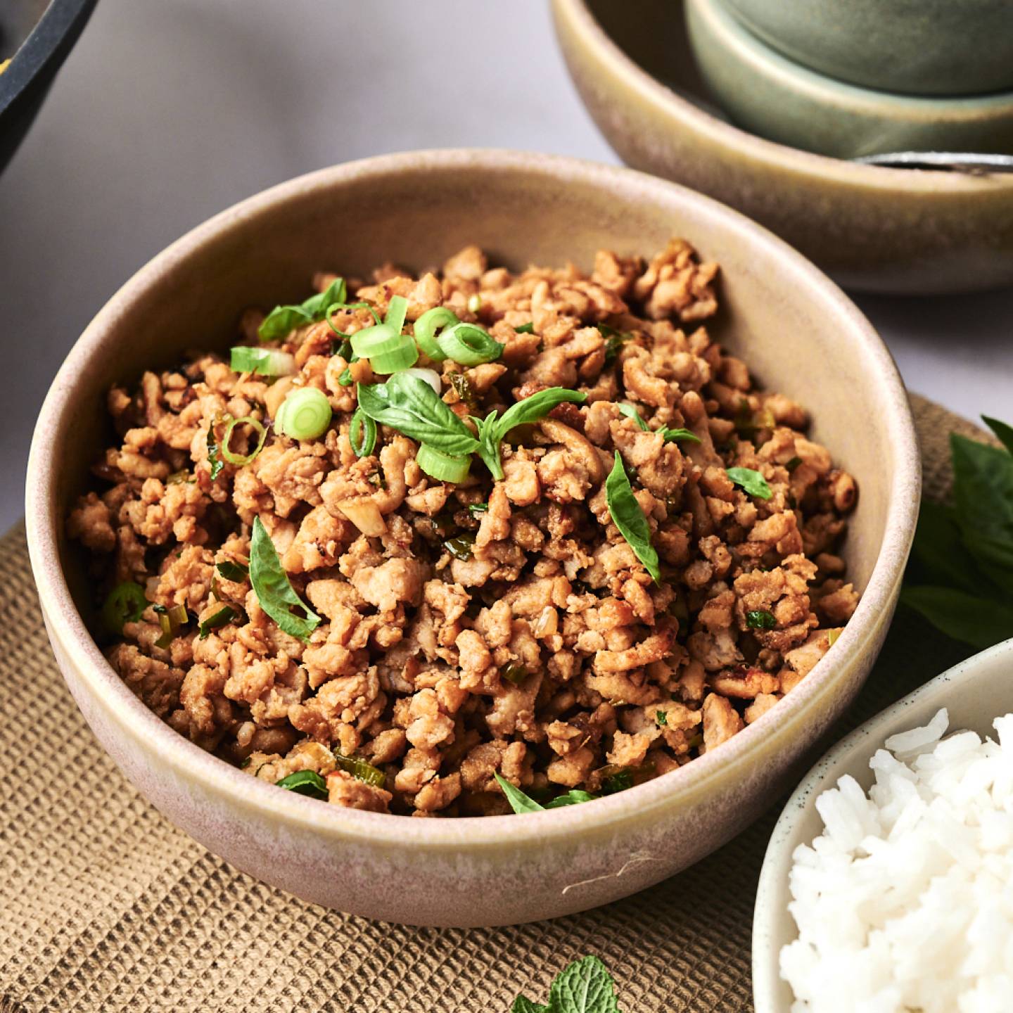 A bowl of seasoned ground meat topped with green onions and basil, served alongside a side of fluffy white rice.