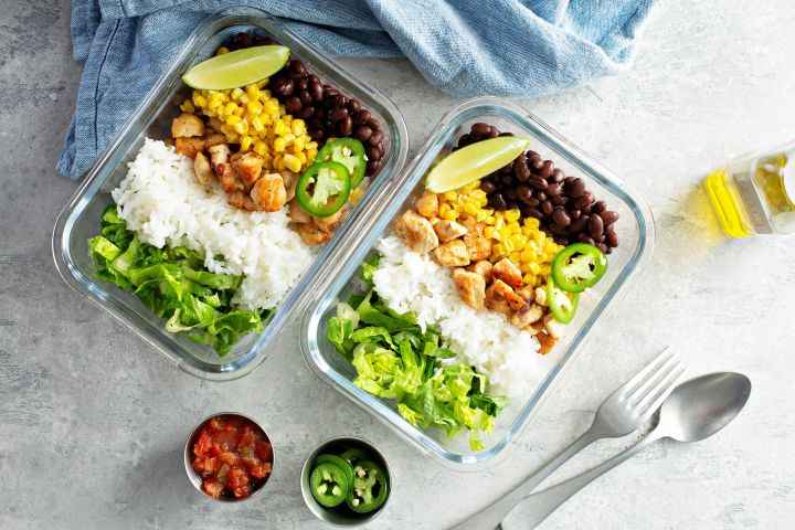 Meal prepped chicken burrito bowls in two glass containers with chicken, rice, beans, corn, and lettuce.