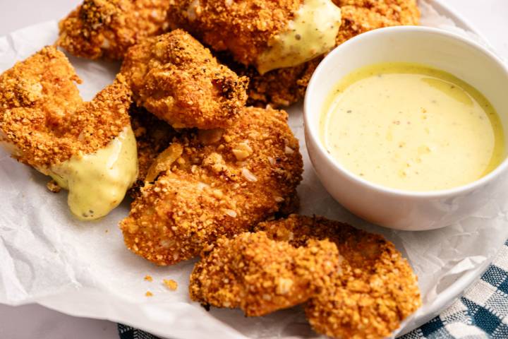 Crispy, golden-brown chicken pieces with mustard coating on a white plate, accompanied by a bowl of creamy lemon-colored dipping sauce.