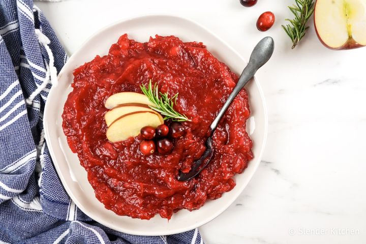 Apple cranberry sauce with fresh cranberries, aplpes, and rosemary in a bowl with a spoon and blue napkin.