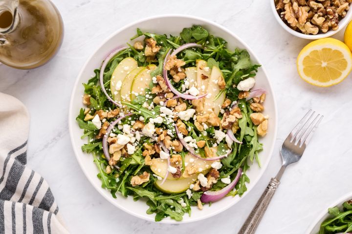 Arugula salad with sliced pears, toasted walnuts, red onions, goat cheese, and vinaigrette tossed in a ceramic bowl.