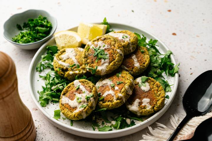 Plate of golden falafel drizzled with creamy sauce, garnished with parsley and lemon wedges. Nearby is a small bowl of chopped herbs.