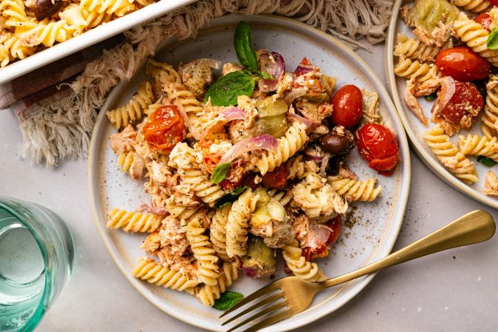 Close-up angled shot of a plated serving of salmon pasta with roasted tomatoes, artichokes, and basil, with a second plate and gold forks partially visible alongside it.