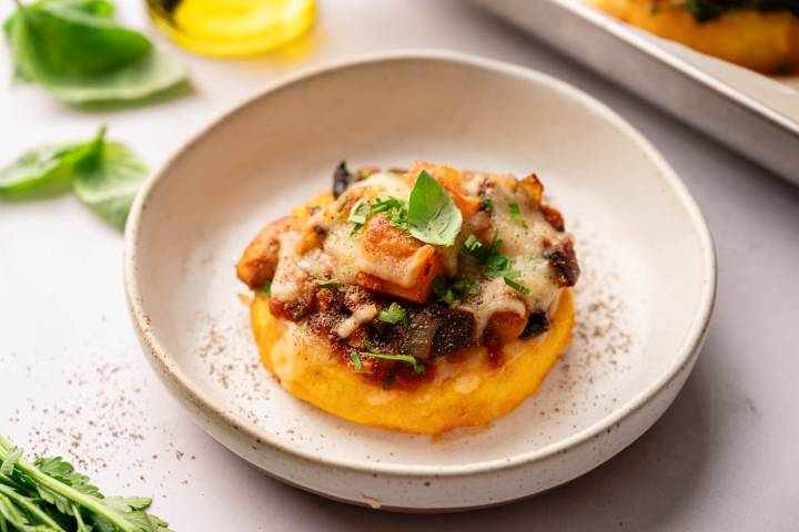 Polenta cake topped with tomato sauce, melted cheese, mushrooms, and herbs on a white plate. Fresh basil leaves and herbs surround the dish.