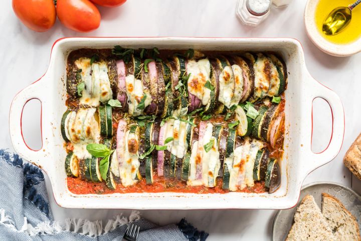 Baked ratatouille with sliced eggplant, zucchini, red onions, and tomatoes in a baking dish with melted cheese and fresh basil.