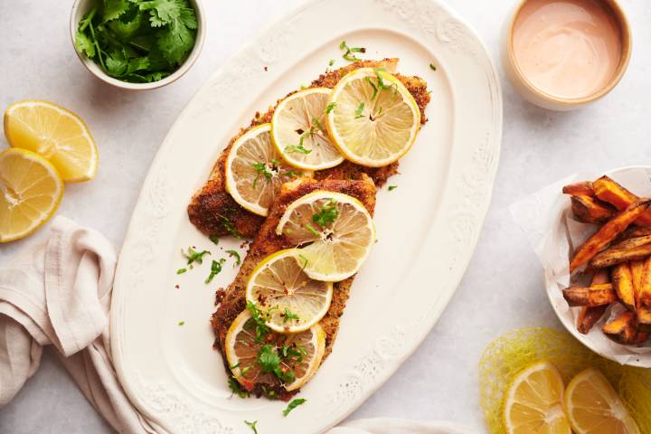 Baked red snapper with spice rub and lemon slices on a plate with sweet potatoes, lemon wedges, and parsley on the side.