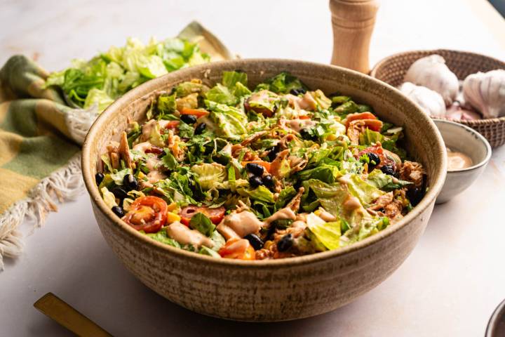 Large serving bowl filled with BBQ chicken chopped salad topped with barbecue ranch dressing, fresh cilantro, black beans, corn, cherry tomatoes, and chopped romaine.