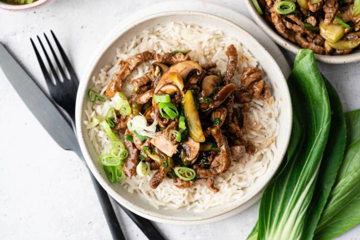 A bowl of rice topped with stir-fried beef, mushrooms, zucchini, and green onions. Nearby are black cutlery and fresh bok choy leaves, suggesting an Asian-inspired meal.