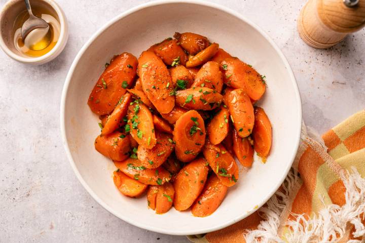 A bowl of glazed carrot slices garnished with parsley, on a white surface. A small bowl with a spoon and a draped cloth are nearby.