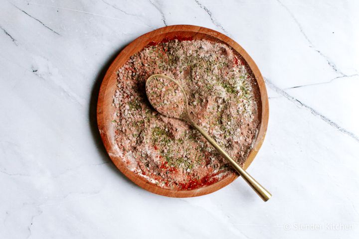 Cajun seasoning blend with smoked paprika, garlic powder, onion powder, oregano, and thyme on a wooden plate with a gold spoon.