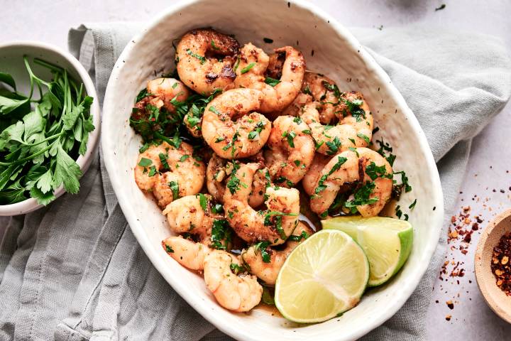 Camarones al ajillo in a white dish with butter garlic sauce, parsley, chile peppers, and lime wedges. 