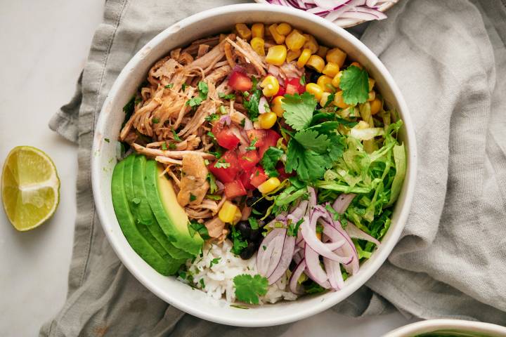 Carnitas burrito bowls with shredded pork, rice, black beans, corn, pico de gallo, lettuce, avocado, and lime wedges.