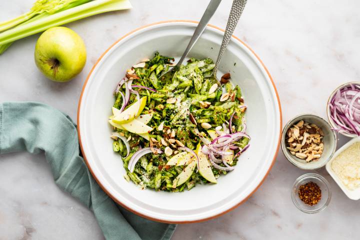 Celery salad with sliced celery stalks, fresh herbs, vinaigrette, apples, almonds, and red onion served in a white bowl.