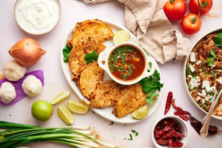 Chicken birria tacos with corn tortillas filled with melted cheese and served with consommé, cilantro, onion, and lime juice.