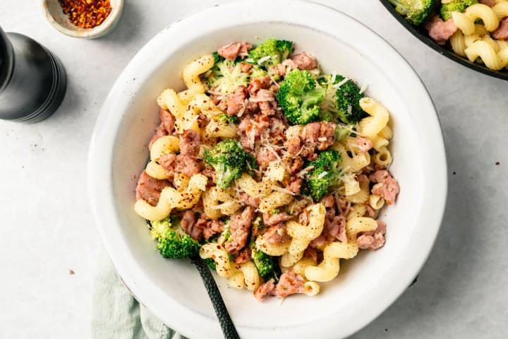 A bowl of curly cavatappi pasta mixed with broccoli and crumbled sausage, topped with grated cheese and black pepper. A black fork rests inside.