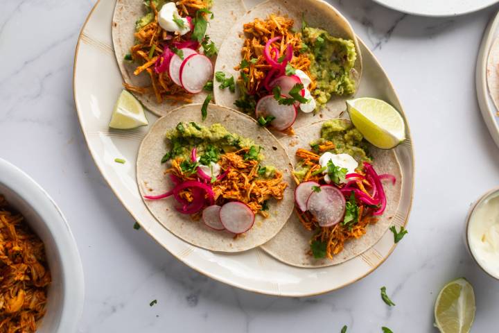 Cochinita pibil tacos with marinated shredded pork, guacamole, cilantro, pickled red onions, and limes in corn tortillas.