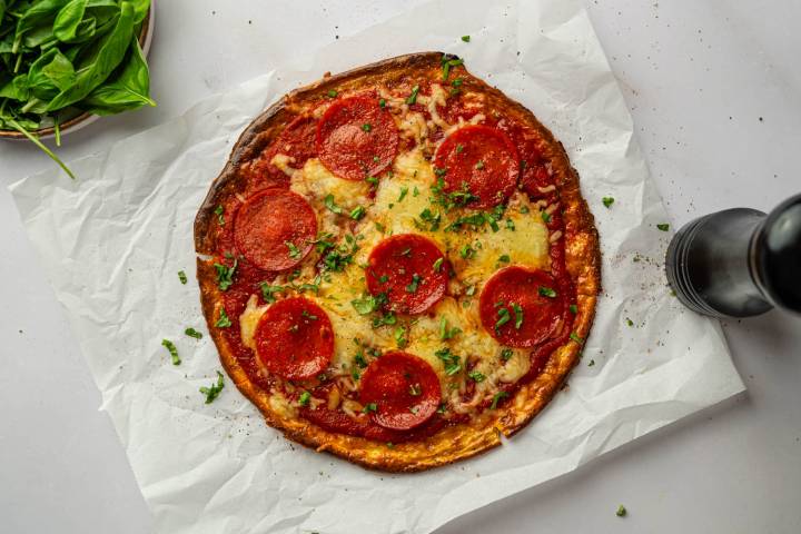 Whole cottage cheese pepperoni pizza with melted mozzarella cheese, turkey pepperoni, and chopped basil on parchment paper, viewed from above.
