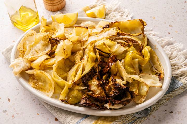 A white plate of roasted cabbage with crispy edges, garnished with lemon wedges. A bottle of olive oil and a wooden pepper mill are in the background.