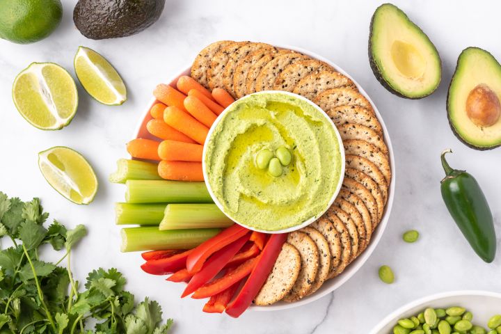 Edamame hummus with frozen edamame, cilantro, avocado, and lime on a plate with vegetables and crackers.