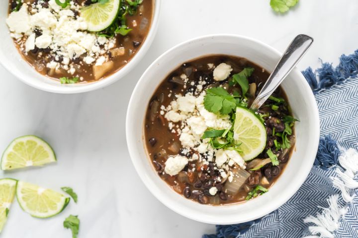 Black bean soup in two bowls with black beans, tomatoes, and onions topped with cilantro, limes, and cheese.