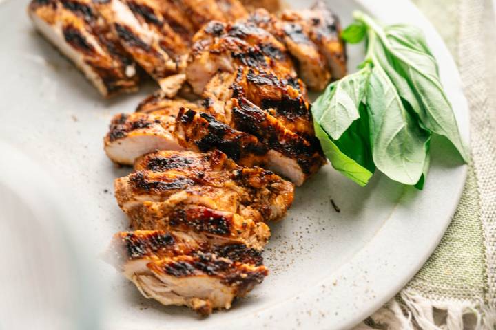 Close-up of juicy grilled chicken breast sliced into pieces, showing charred grill marks and a tender interior, plated with fresh basil leaves.