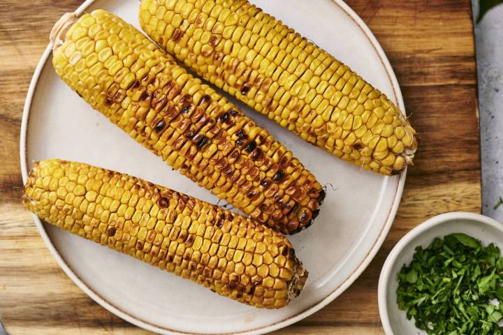 Three ears of grilled corn with lightly charred kernels arranged on a white plate, with a small bowl of chopped fresh cilantro on a wooden cutting board.