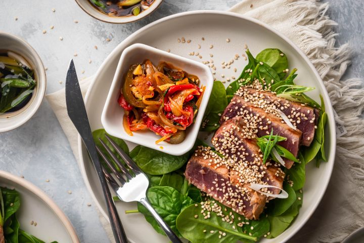 Grilled tuna with soys sauce and sesame seeds on a plate with green onions, lettuce, and cooked peppers.