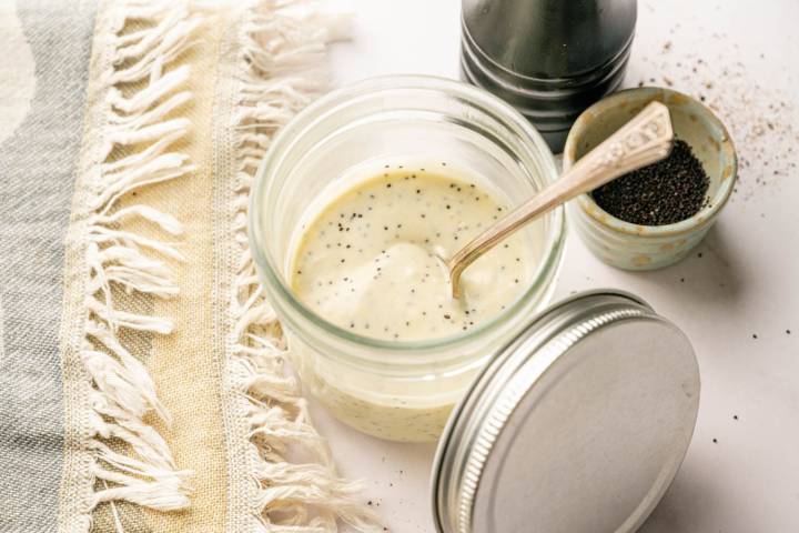 Jar of healthy poppy seed dressing with black pepper and poppy seeds nearby, styled on a neutral kitchen surface with a linen towel.