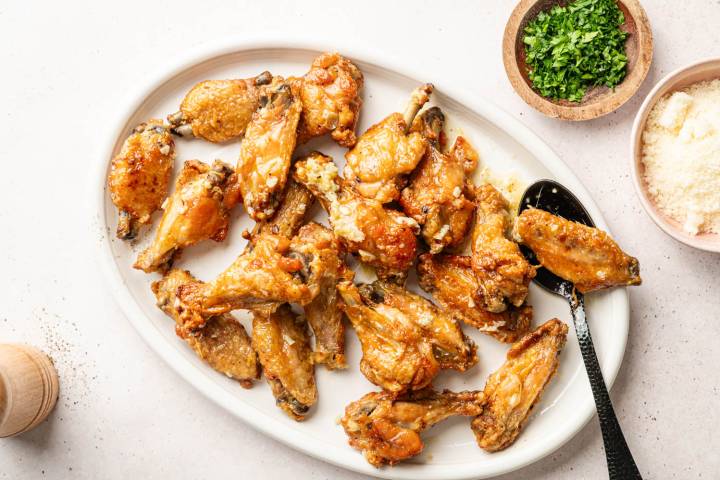 Golden-brown chicken wings on an oval white plate, garnished with garlic. Bowls of chopped parsley and grated cheese are beside the dish.