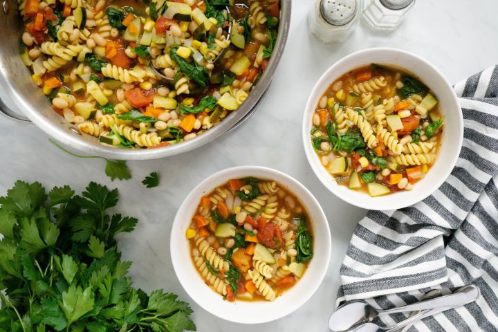 Minestrone soup with zucchini, yellow squash, pasta, white beans, spinach, and tomatoes in two bowls. 