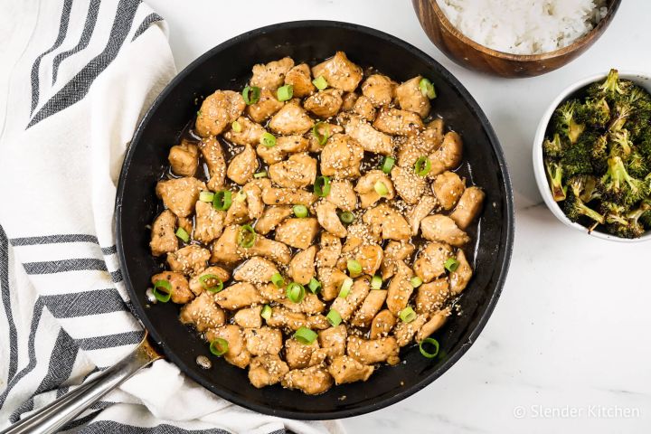 Healthy Sesame Chicken in a skillet with green onions with white rice and broccoli on the side.