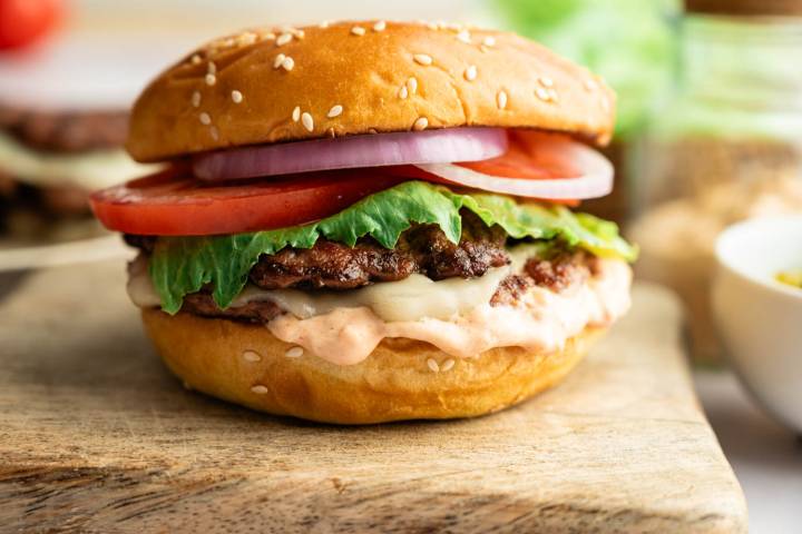 Side view of a homemade smash burger with melted cheese, crisp lettuce, sliced tomato, red onion, and burger sauce on a toasted brioche bun.