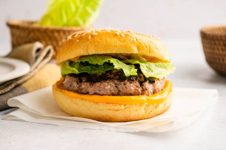 Close-up of a finished turkey burger on a sesame seed bun with melted cheese and crisp lettuce, set on parchment paper.