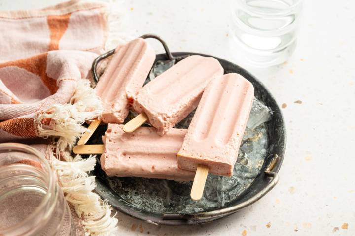 Four homemade high-protein Greek yogurt popsicles resting on ice in a metal serving tray with a striped kitchen towel nearby.