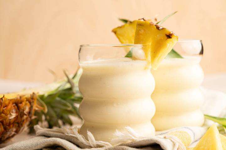 Two tropical protein smoothies topped with pineapple slices, photographed on a light background with fresh pineapple pieces and leaves scattered around.