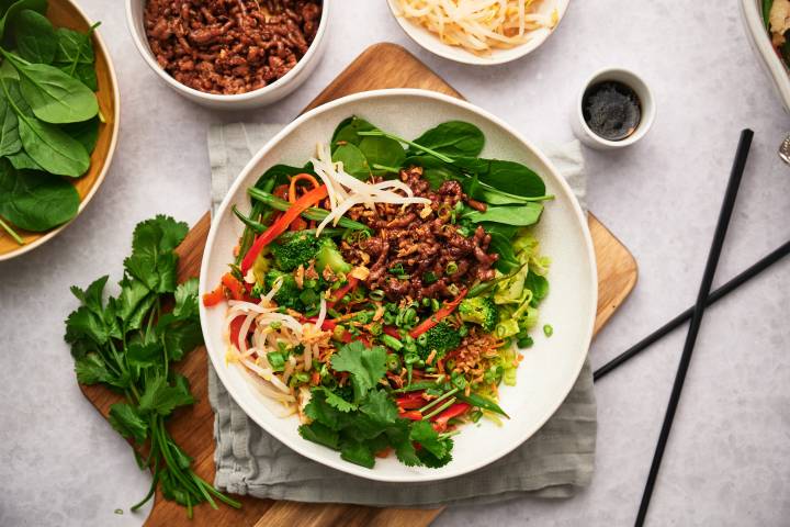 Korean beef bowls with rice, cilantro, carrots, bell peppers, and sprouts in a bowl with spicy sauce. 