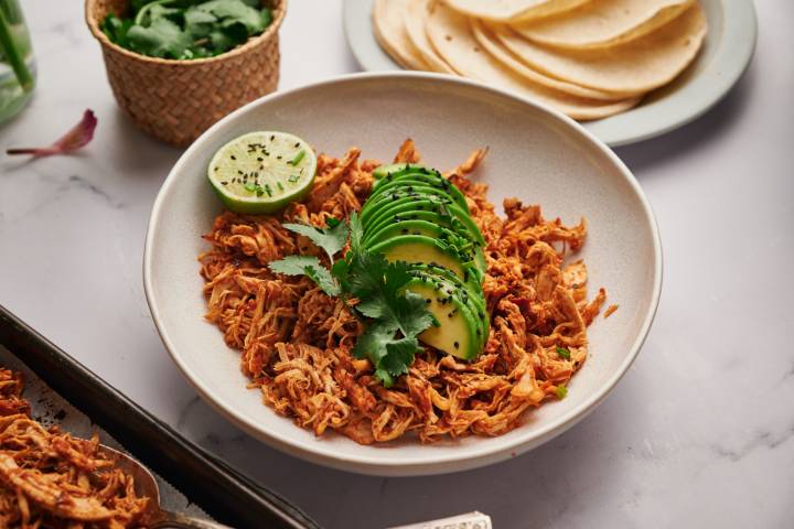 Mexican shredded chicken with tomatoes, spices, and chipotle peppers in a bowl with avocado, lime, and corn tortillas.