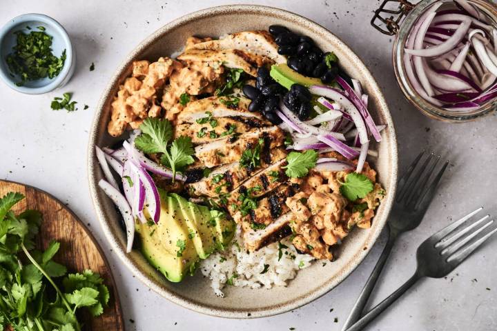 A bowl filled with grilled chicken, rice, black beans, avocado, red onion, and creamy sauce, garnished with fresh cilantro.